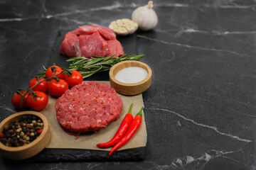 Close-up on the left is a raw beef round patty lying on paper surrounded by seasonings and vegetables, behind them separated by a sprig of rosemary lies meat cut into chunks on a dark background with