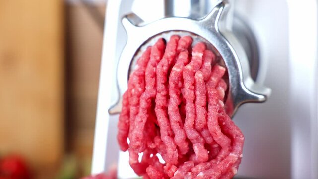 A close-up side view of a metal meat grinder, from which red minced meat from pork and beef comes out against the background of wood and vegetables. The concept of cooking, healthy food, dietary and