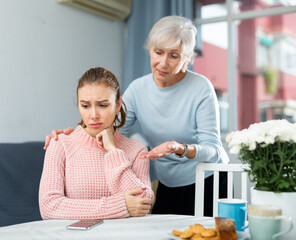 Upset young woman sitting at table. Her mother senior woman and trying to cheer her up.