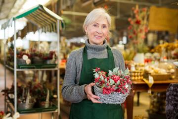 Positive mature woman in apron holding christmas decoration, standing in store, smiling and looking at camera.
