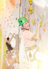 Obraz premium Side view of sportive mature female climber moving up, using safety top rope and climbing artificial wall indoors. Extreme sports and bouldering concept