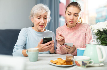 Positive senior woman and her adult daughter sitting at table in cozy dining room, looking...