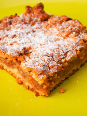 A yellow plate with a piece of cake on it. The cake is covered in powdered sugar. The plate is placed on a white background. Premium slice of a fine bakery product