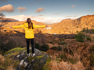 Fototapeta premium Teenager girl looking at stunning nature scenery from Ladies viewpoint in Killarney, Ireland. Travel and tourism concept. Model wearing yellow jacket and has long hair. Warm sunny day.