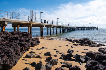 Cowes Jetty extends over the calm waters of Western Port Bay, with tourists walking along it. It is...