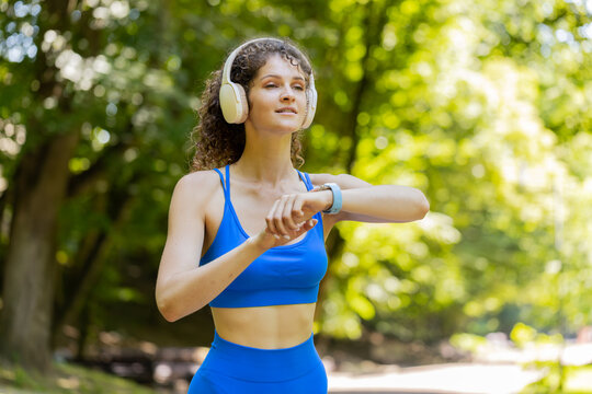 Athletic runner adult girl looks at smart watch device fitness bracelet checking time, heart rate, pulse, SPO2 on smartwatch on jogging. Young woman working out cardio outside in city park in morning.