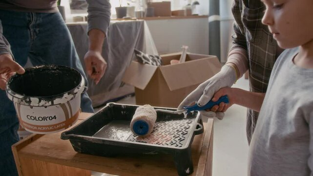 Young man pouring grey paint from plastic bucket into tray while his wife and daughter with paintroller mixing it and preparing for painting walls in living room during renovation