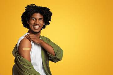 Vaccination Concept. Cheerful Black Guy Demonstrating Arm With Adhesive Bandage After Covid-19 Vaccine Shot, Happy African Man Protecting Himself From Coronavirus, Posing Over Beige Background
