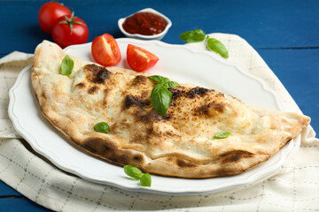Tasty calzone with basil, tomatoes and sauce on blue wooden table, closeup