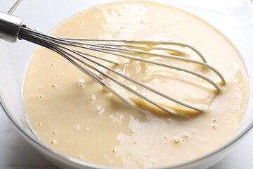 Whisk and bowl of dough on white table, closeup