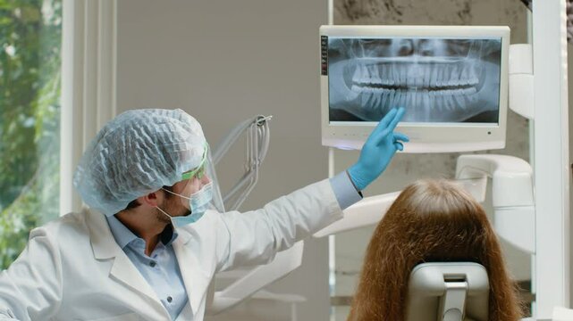 A dentist in a white coat points at a dental x-ray on a monitor while discussing treatment with a patient in a modern dental clinic. The patient is seated, looking attentively at the screen.
