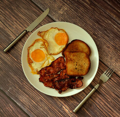 A plate of delicious breakfast, scrambled eggs, wheat toast and slices of fried bacon on a wooden table, with cutlery nearby.