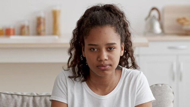 A teenage girl sits alone on a couch in her cozy living room, deep in thought. The bright space features a coffee table and shelves with various items, capturing a moment of reflection.