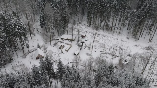 A snow-covered village of wooden houses lies deep in the wilderness. The drone view reveals a cluster of rustic dwellings surrounded by a dense winter forest, untouched by modern times.