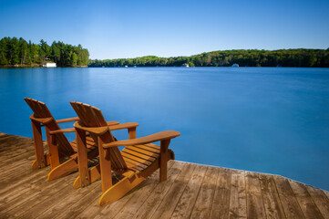 Two Muskoka chairs sitting on a wood dock facing a lake. Across the calm water are cottages nestled among green trees. Long exposure capture.