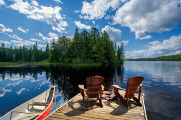 Two Adirondack chairs on a wooden dock overlook the blue waters of a Muskoka lake. A life jacket and paddles are visible, while a red canoe is tied to the dock.