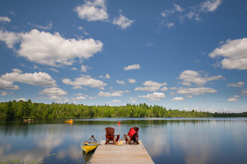 Two Adirondack chairs on a wooden dock overlook the blue waters of a Muskoka lake. A life jacket...