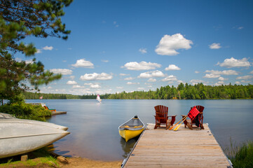 Two Adirondack chairs on a wooden dock overlook the calm blue waters of a Muskoka lake. A life...