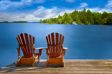 Two Adirondack chairs sitting on a wooden dock facing a blue calm lake. Across the water is a white...