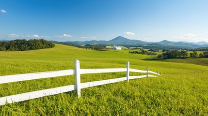 Fototapeta premium White wooden fence stretches across a lush green rolling meadow under a clear blue sky with distant mountains.