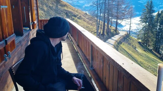 A man sits on a wooden chalet balcony, smoking and gazing thoughtfully at the valley of Simmental in Switzerland, enjoying the fresh air and scenic view on a crisp, sunny spring morning