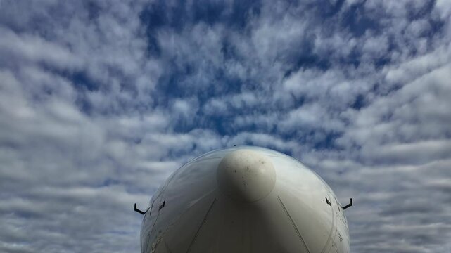 External view, extreme low angle shot, of the nose of a white color fudelage jet at the stand under a blue sky with some passing fluffy clouds. Timelapse. 4K