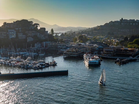 Aerial view of a sailboat navigating Tiburon harbor, with docked yachts and a ferry. Coastal town nestled on hillsides. Belvedere Tiburon, San Francisco, California, USA