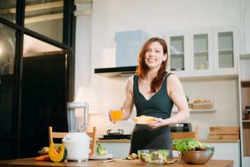 Smiling woman in a modern kitchen with fresh fruits, veggies, blender, and juice, embracing clean...