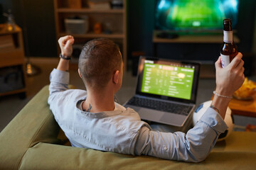 Person sitting on couch watching soccer game on laptop, with hand holding beer bottle, providing a casual and relaxed atmosphere