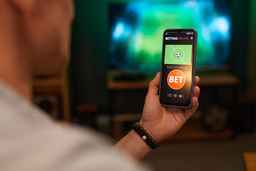 Person checking and placing bet on soccer match using smartphone while relaxing at home, displaying betting platform on screen
