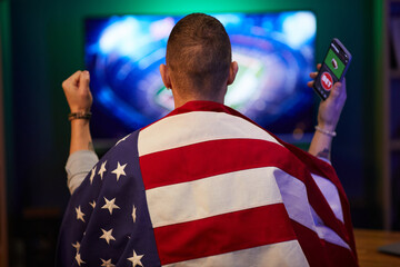 Person draped in USA flag seen from behind, celebrating gaming victory with clenched fist and mobile phone, showing pride and enthusiasm in energetic scene