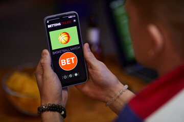 Person holding smartphone displaying online betting application in relaxed indoor setting Hand showing screen with basketball game options and bet button