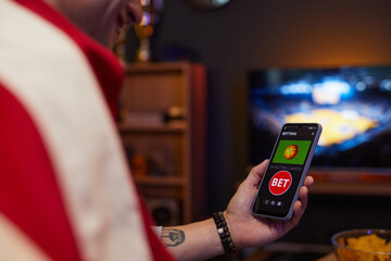 Person betting on basketball match using betting app on smartphone while watching game on television in dimly lit room with cozy atmosphere