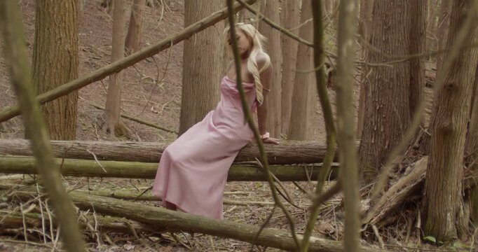 Young ebony woman with long blonde hair, sits on top of fallen tree trunks in the woods. She's wearing a pink gown, and scrunchie on her right wrist. Wide shot, handheld camera tracking side to side.