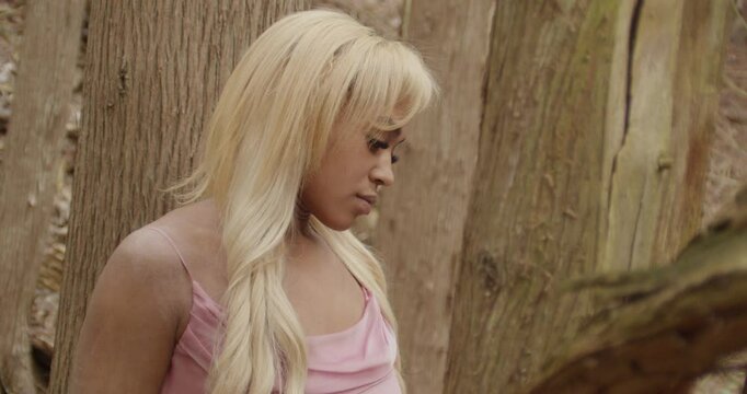 Young ebony woman wearing a pink gown. She sits alone in the forest, surrounded by trees. She shifts her gaze from a distance, to camera, then looks up to the sky. Slow Motion 60fps. Handheld camera.