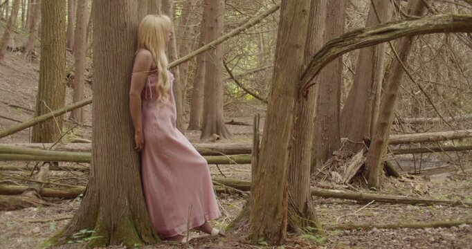 Young ebony woman leans against the trunk of a sweetgum tree, in the forest, She is relaxed, enjoying the sights surrounding her. She has long wavy blonde hair, wearing a pink gown, 60fps slow motion.