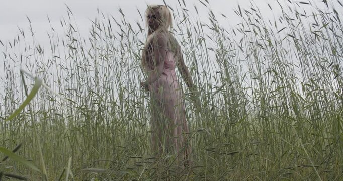 Young ebony woman in a pink silk satin dress, with long blonde hair; standing in a field of tall grass. She runs her hands along the swaying tall grass. Wide shot, Handheld camera tracking both sides.