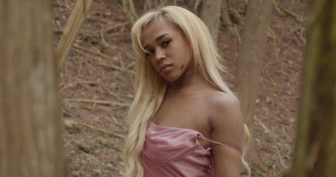 Young black woman wearing a pink gown. She sits alone in forest of trees, seductively gazing at the camera while playing with her blonde hair. Scrunchie on right wrist. Medium Shot Slow Motion 60fps.