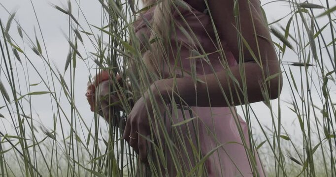 Young ebony woman, with long blonde hair. She is wearing a pink satin gown, with a matching scrunchie on her right wrist. She runs the tall grass between her fingers. 60fps slow motion. Camera Tilts.