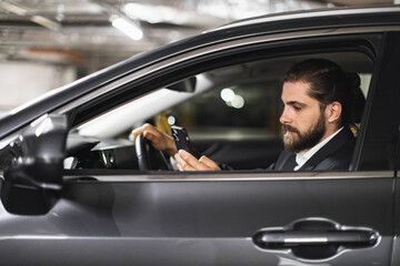 Caucasian male in suit using smartphone inside car parked indoors. Young adult appears focused on device, emphasizing concept of distracted driving.