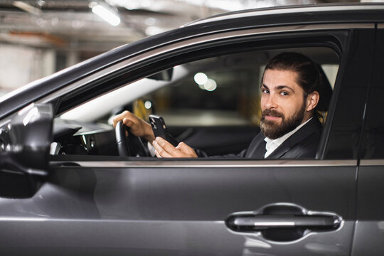 Caucasian adult man driving car using smartphone. Business professional wearing suit looking confident. Interior car shot in parking garage.