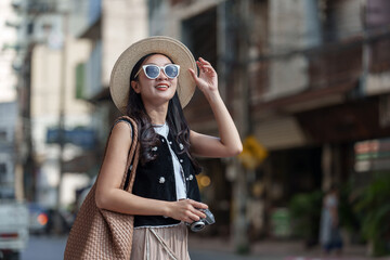 Young woman tourist wearing a stylish hat and sunglasses, holding her hat and camera, happily...