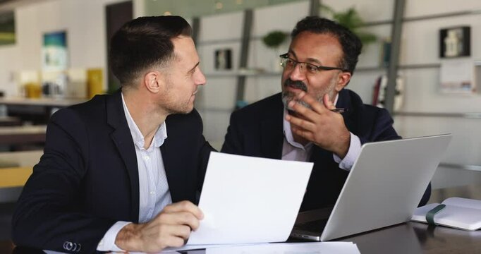 Positive multiethnic male co-workers working on project together, talking at laptop, discussing sales report, business plan, Internet marketing, paper document, enjoying teamwork