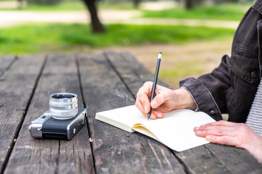 Female journalist taking notes on a notebook with a pencil at a wooden table outdoors in a park, with a vintage camera lying on the table next to her