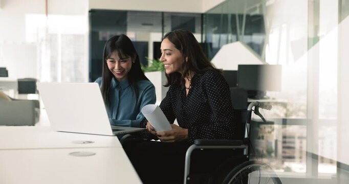 Happy multiethnic project coworkers discussing online software product at laptop, pointing at screen, speaking, smiling, brainstorming. Two employees from inclusive company staff working in office