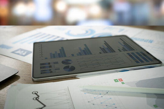 A tablet displaying graphs analyzing sales data and growth graph and charts alongside printed reports on a table.