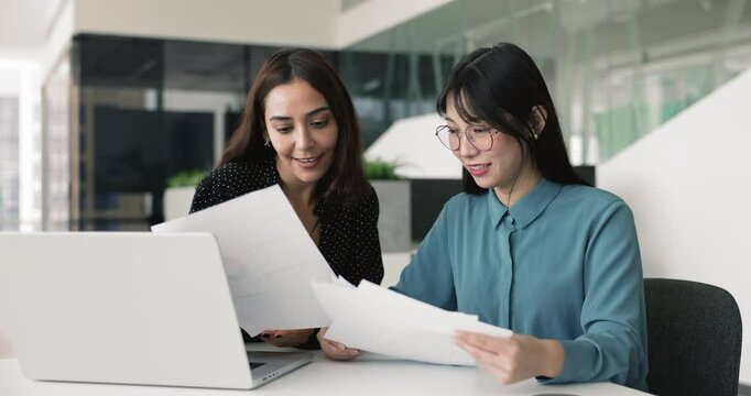 Two positive diverse female coworkers analyzing paper marketing report, studying financial statistic data in documents together, discussing product management, strategy