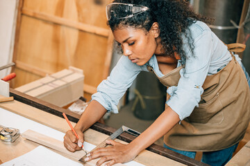 African American carpenter woman drawing a line on wood with a pencil and ruler in workshop. Focused on precise measurements, woodworking, and construction tasks in carpentry, National Carpenters Day