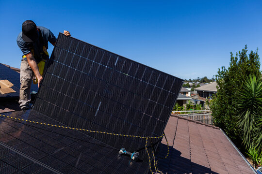 Solar panel technician holding up solar panel on roof during installation in suburbia