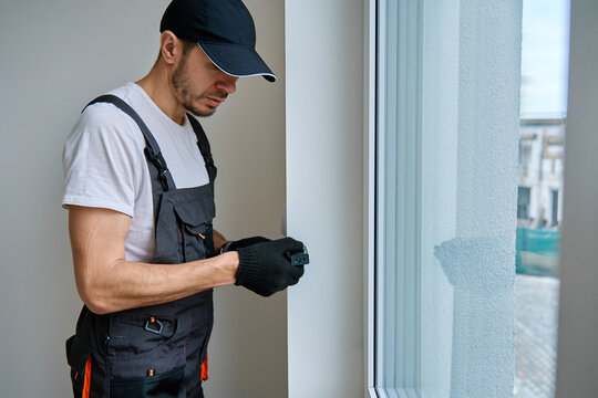 Construction worker in gloves uses laser distance measurer to inspect wall. Builder using digital tool for precision measurement during repair work. Construction and renovation concept